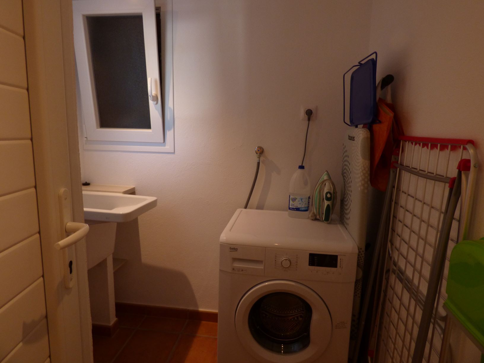 Pieni ja käytännöllinen pyykinhuone, jossa on pyykinpesukone, pieni pesuallas ja ikkuna.