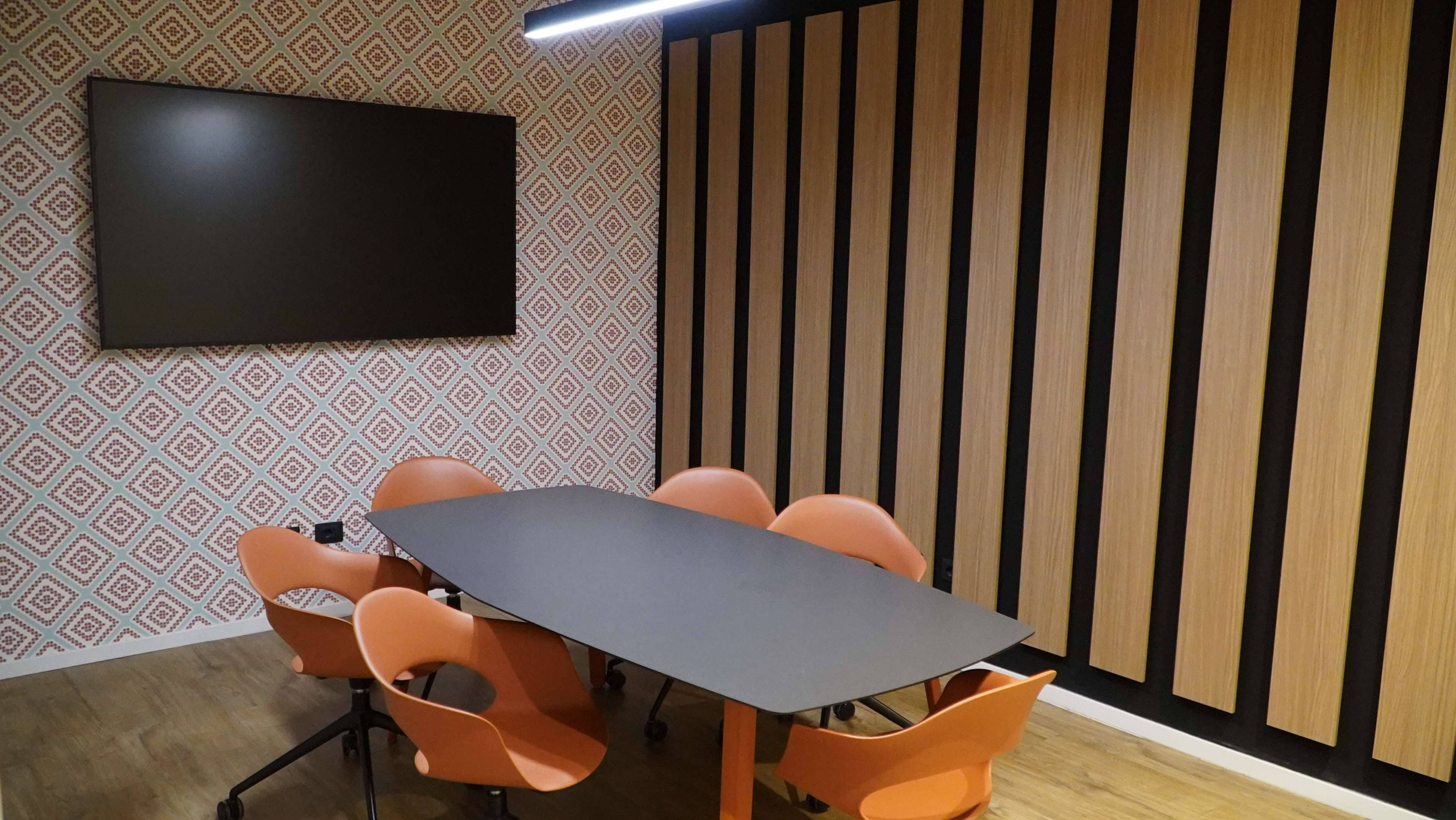 A conference room with a long table, six orange chairs, a flat-screen TV on the wall, and a patterned wallpaper.