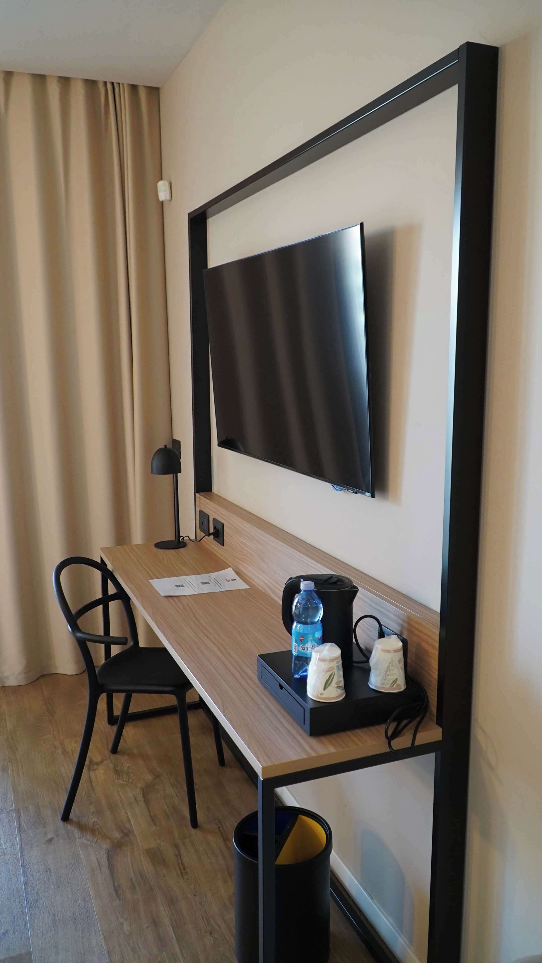 Habitación de hotel con un escritorio de madera, una silla negra, un televisor de pantalla plana montado en la pared, una lámpara negra, una cafetera, una botella de agua y un bote de basura.