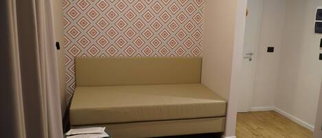 A small, beige sofa in a corner of a room with patterned wallpaper and wooden flooring.