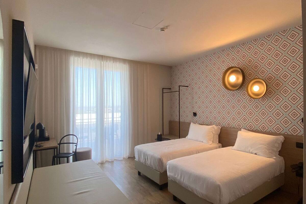 Habitación de hotel con dos camas, un escritorio, una silla y un ventanal con cortinas.