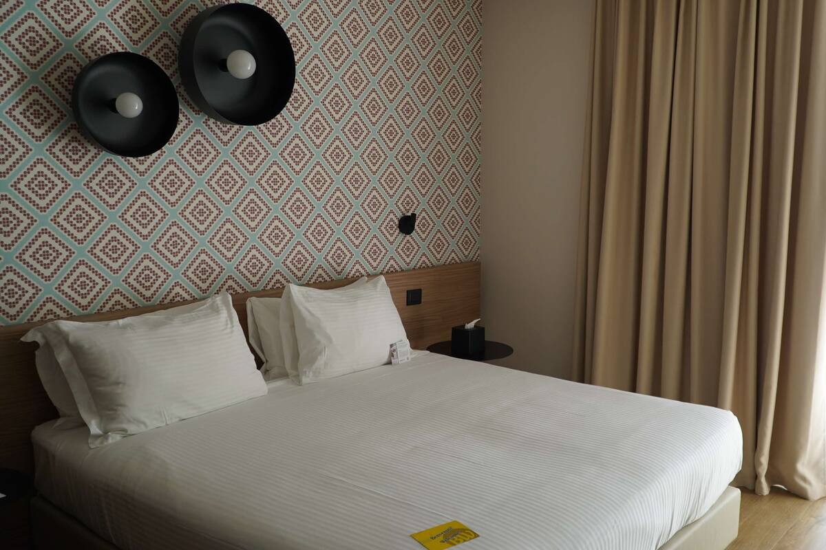 Habitación de hotel con cama, dos altavoces montados en la pared, mesita de noche con caja de pañuelos y papel tapiz estampado.