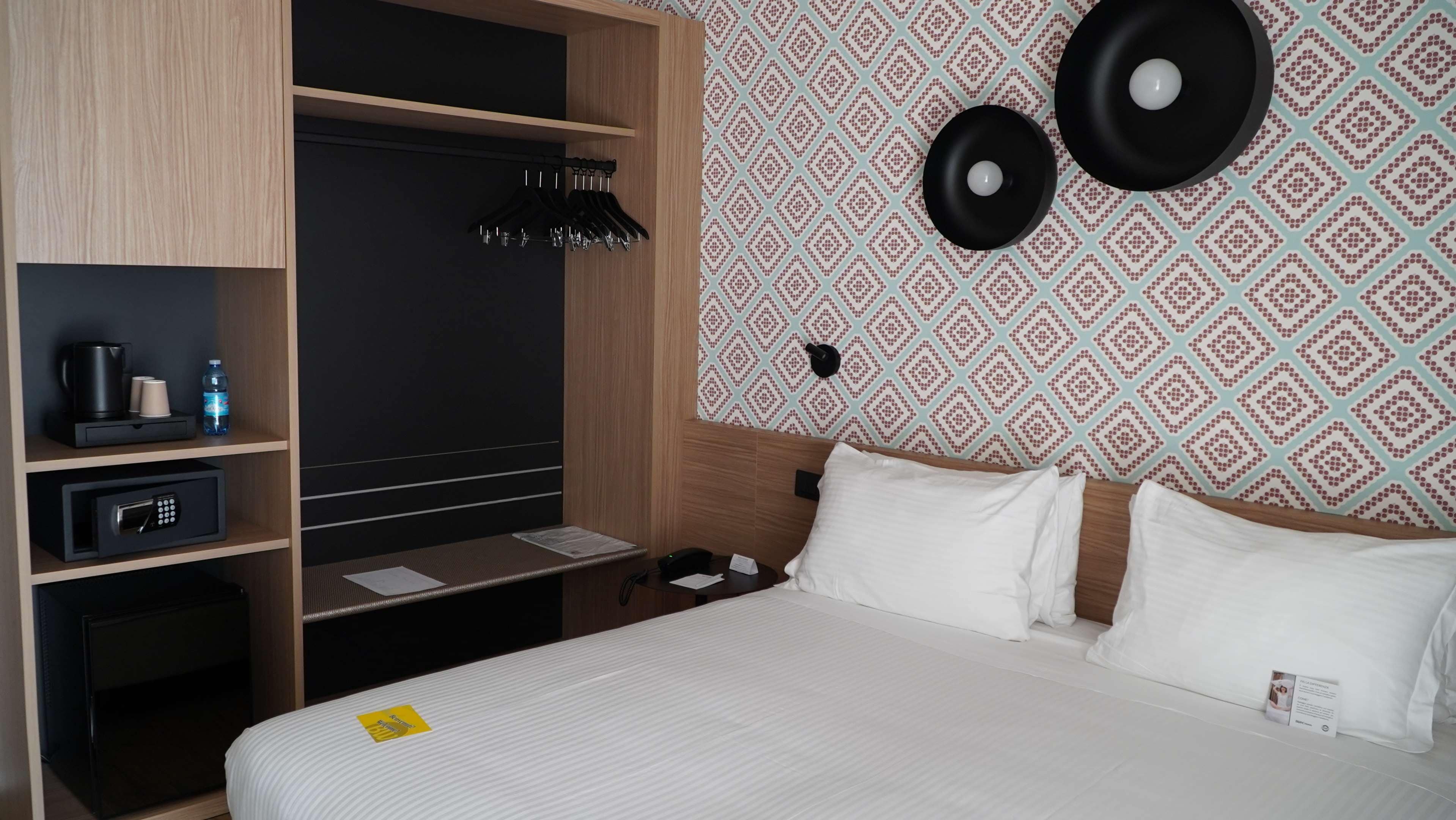 A hotel room with a bed, a desk with a computer, a wardrobe, and a wall with patterned wallpaper.