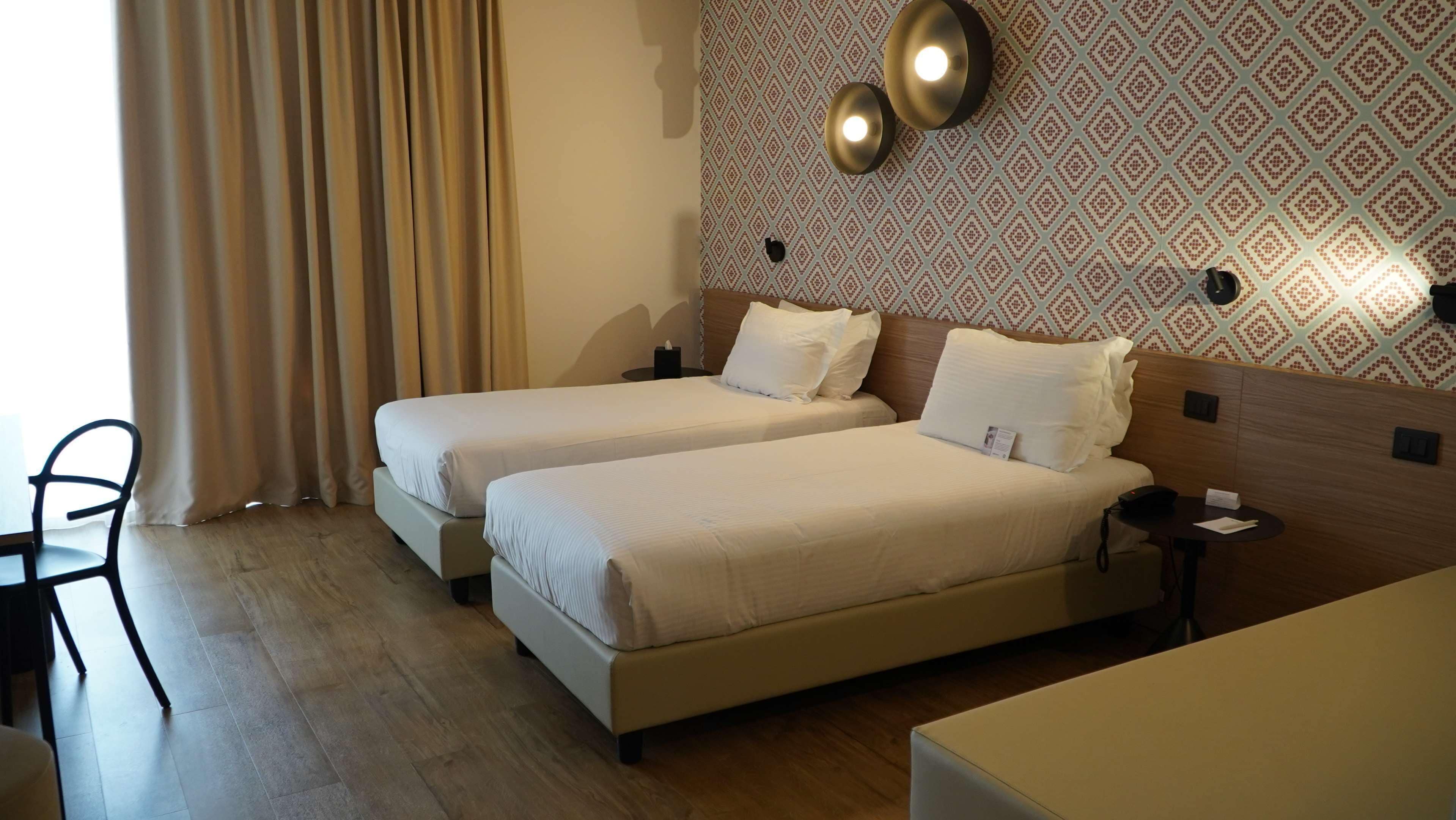 A hotel room with two beds, a desk, a chair, and a patterned wall.