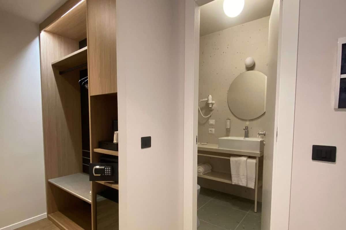 Habitación con piso de madera, armario empotrado con estantes, y baño con espejo, lavamanos y barra para toallas.