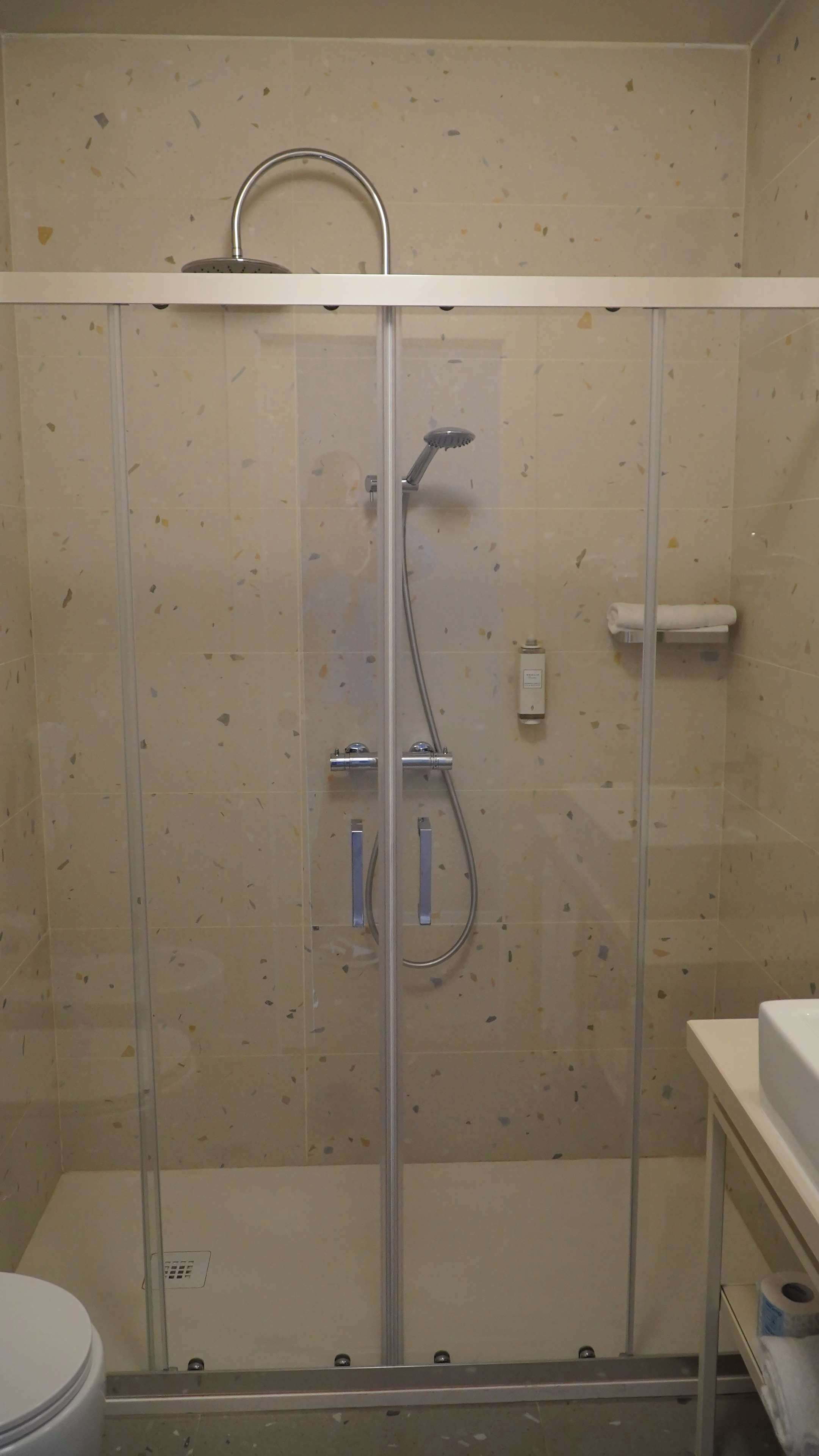 Ducha envuelta en vidrio con un cabezal de ducha de mano, panel de control en la pared y un perchero para toallas.