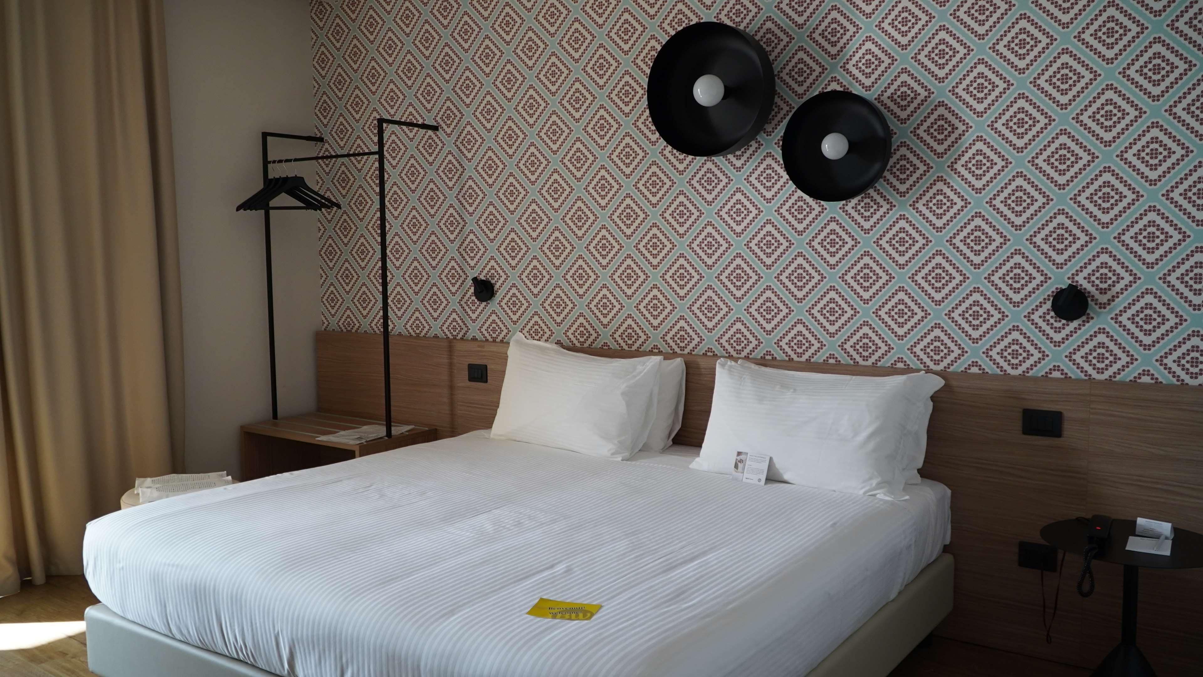 A hotel room with a neatly made bed, a bedside table, a lamp, and a wall with patterned wallpaper.