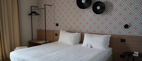 A hotel room with a neatly made bed, a bedside table, a lamp, and a wall with patterned wallpaper.