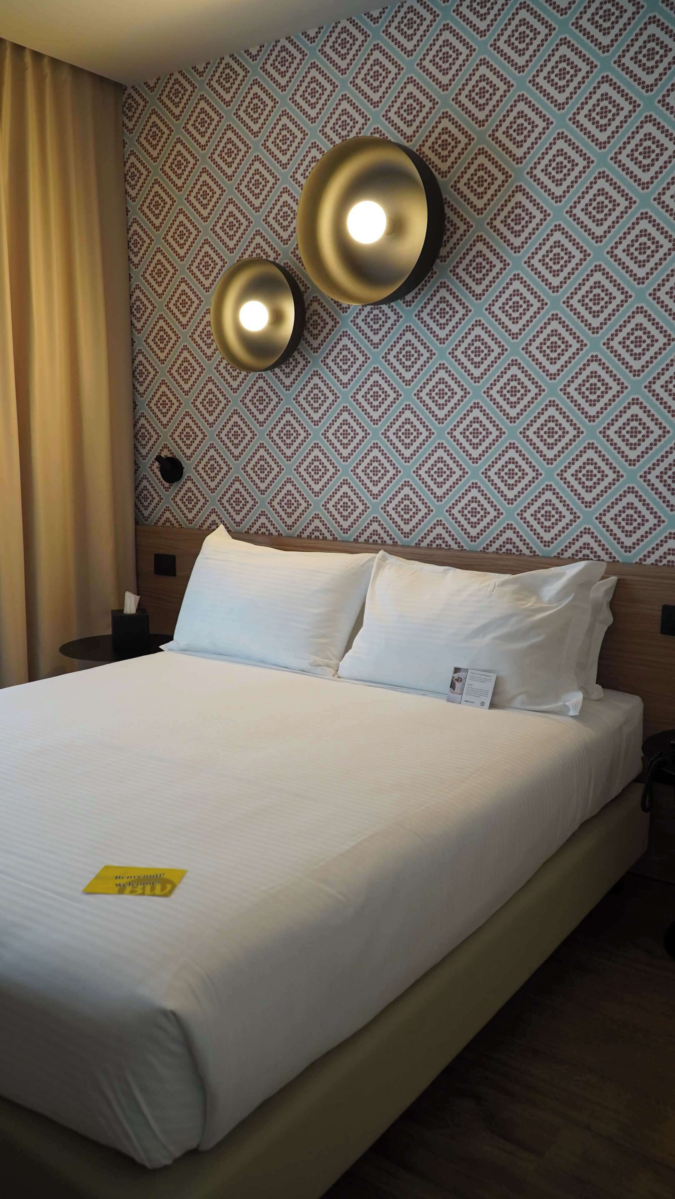 Een net opgemaakt bed met witte lakens, een nachtkastje met een lamp, en een behang met patroon.