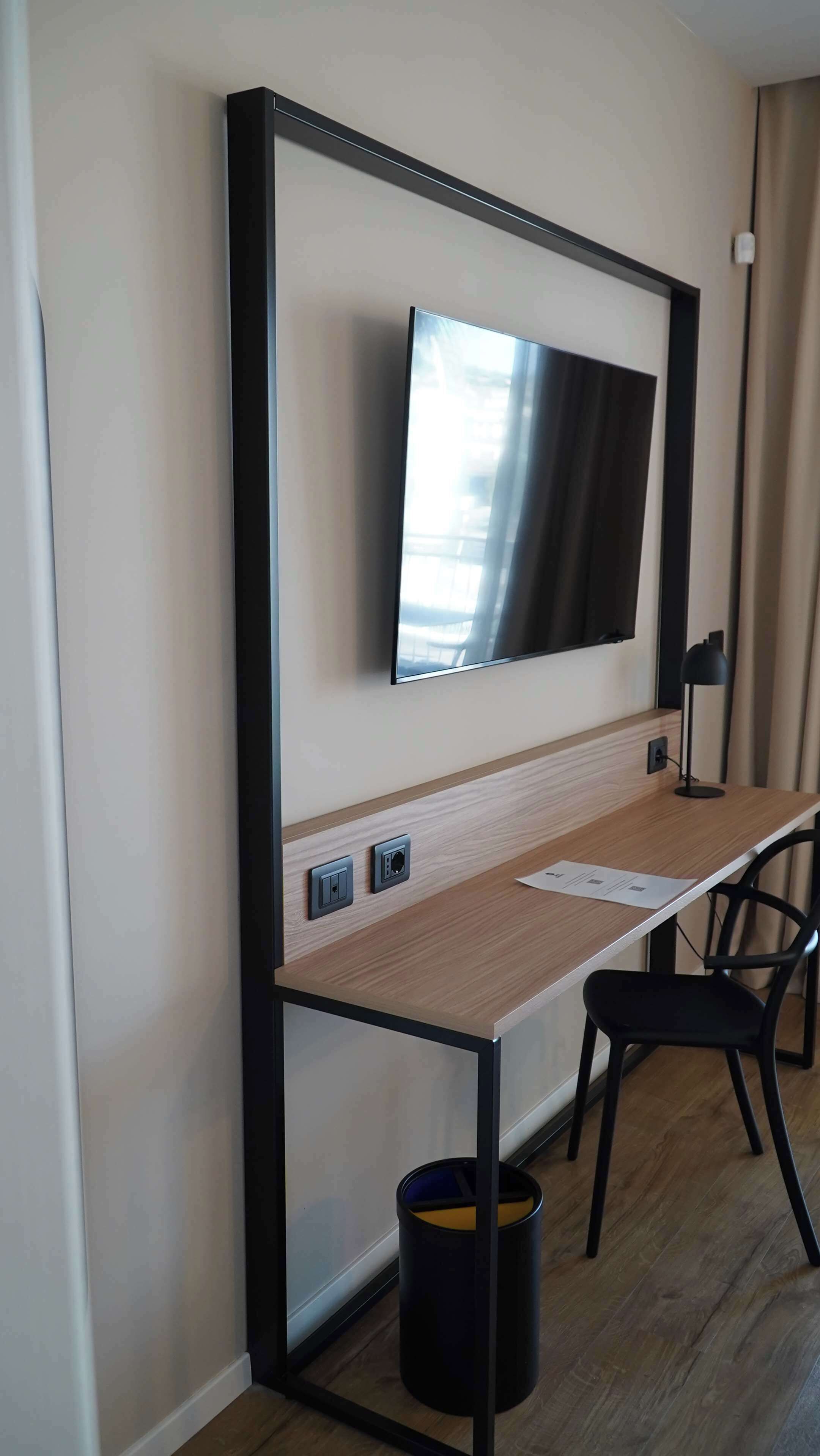 A modern hotel room with a large flat-screen TV mounted on the wall, a wooden desk with a chair, a black trash bin, and a lamp.