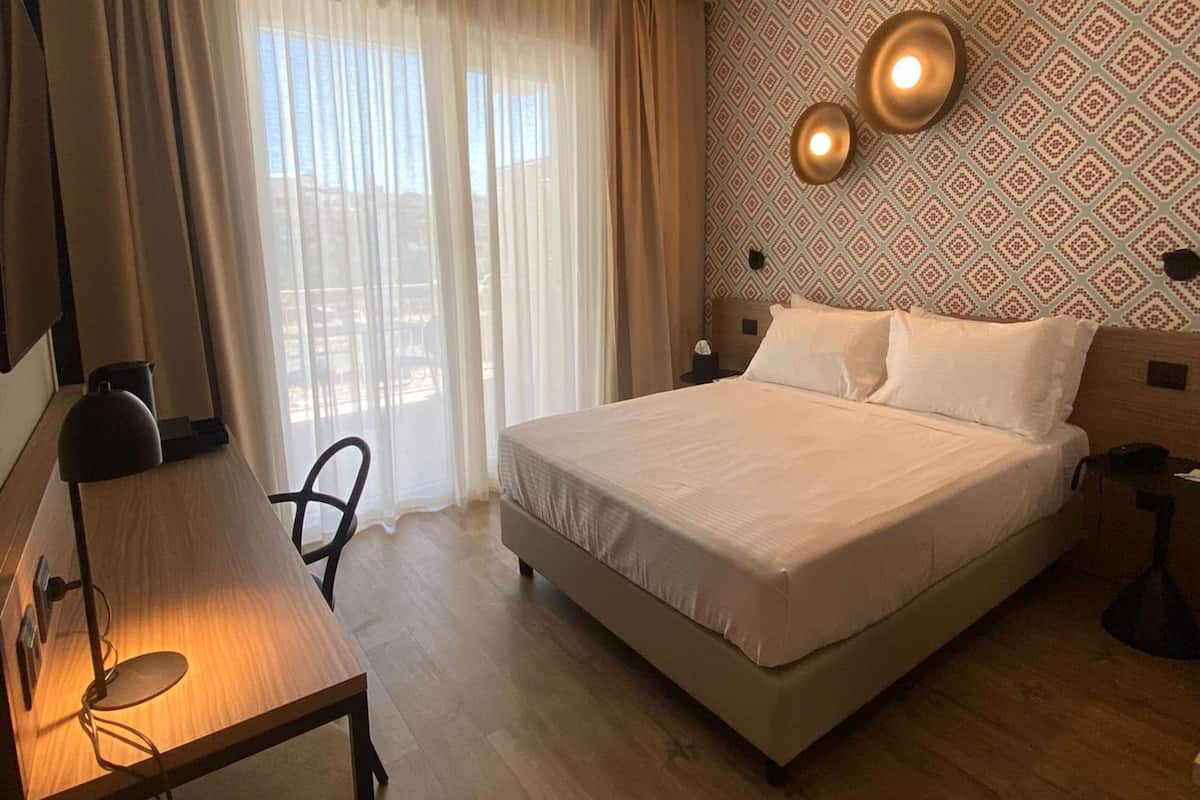 Una habitación de hotel con cama, escritorio con lámpara, silla y ventana con cortinas.