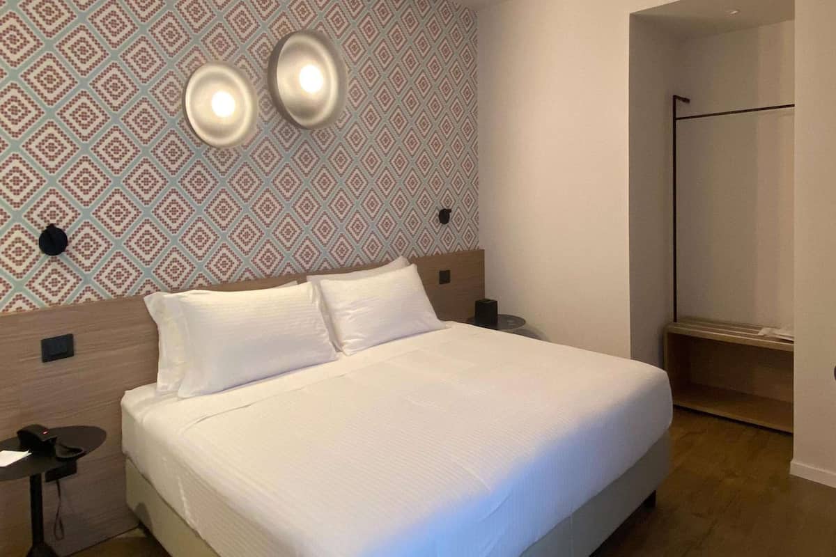 Una cama bien hecha en una habitación de hotel, con papel tapiz estampado, una mesita de noche con lámpara y suelo de madera.