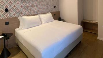 Una cama bien hecha en una habitación de hotel, con papel tapiz estampado, una mesita de noche con lámpara y suelo de madera.