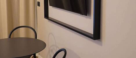 Una habitación con una mesa redonda de comedor y dos sillas, un televisor de pantalla plana montado en la pared y una ventana con cortinas color beige.