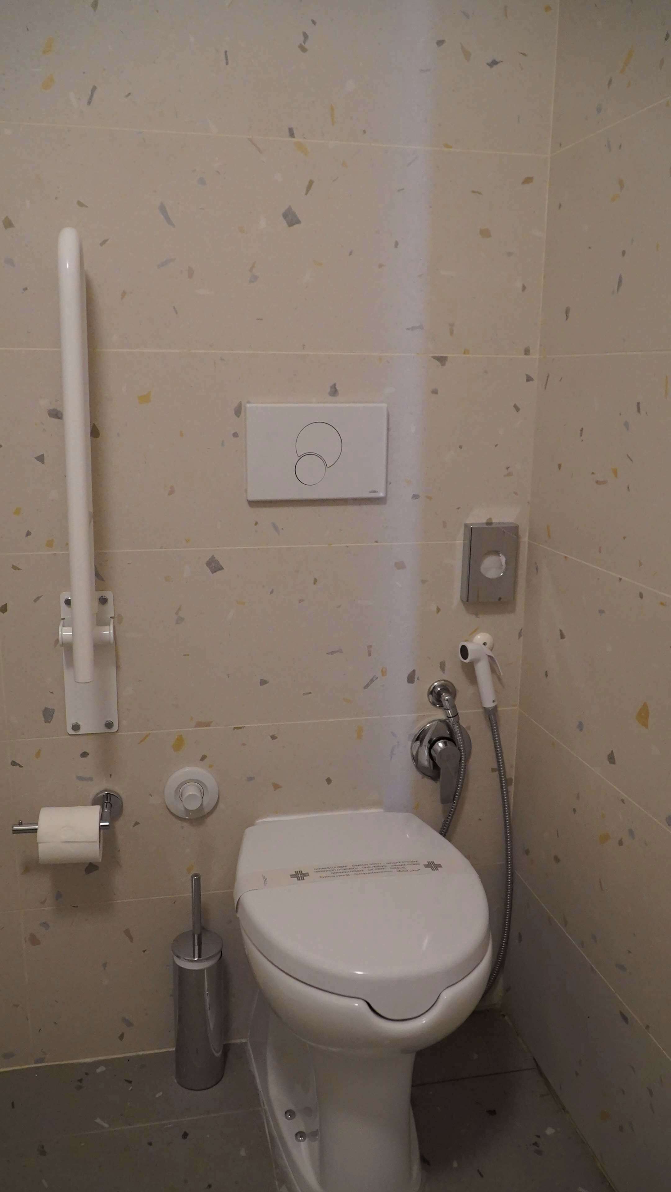 Une petite salle de bain carrelée, équipée de toilettes, d’un sèche-mains, d’un distributeur d’essuie-tout et d’un distributeur de savon.