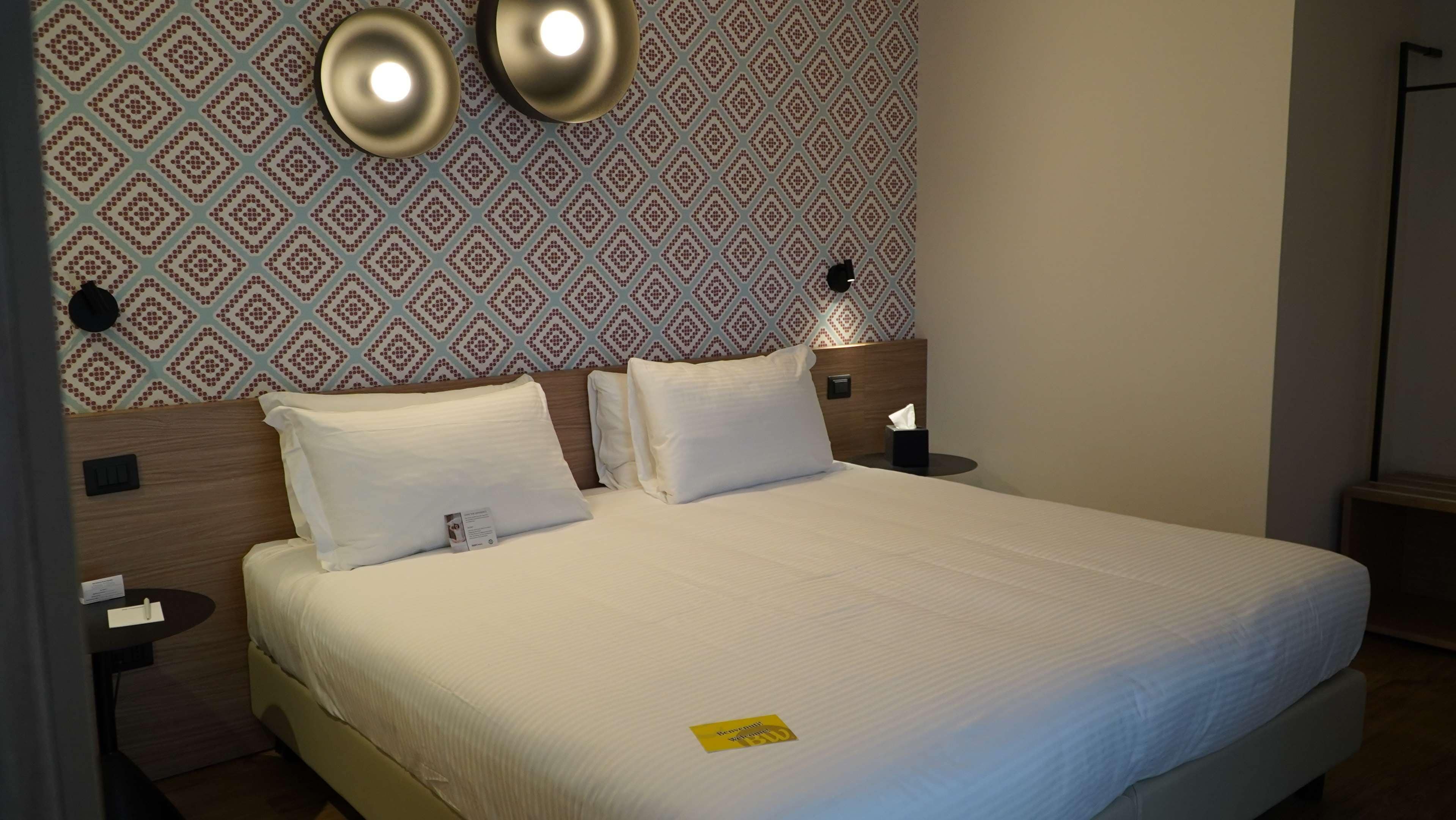 A neatly made bed with white linens, a patterned wallpaper, and two bedside lamps.