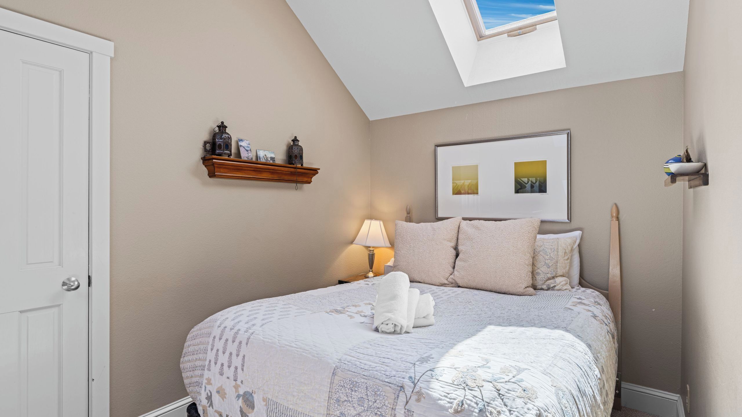 Un dormitorio con tragaluz, una cama con edredón y almohadas blancas, una mesita de noche con lámpara y una estantería con objetos decorativos.