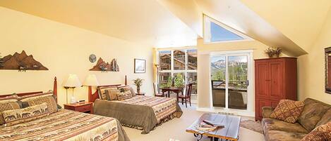 A spacious room with two beds, a sofa, a table, and a view of mountains through large windows.