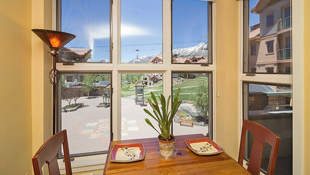 Uma sala de jantar com mesa, cadeiras, um vaso de planta e vista para a rua e edifícios através de grandes janelas.
