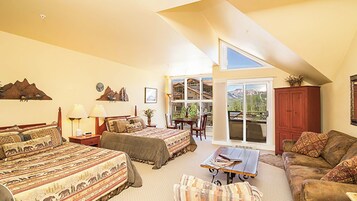 Quarto com duas camas, um sofá, uma mesa e vista para montanhas por grandes janelas.