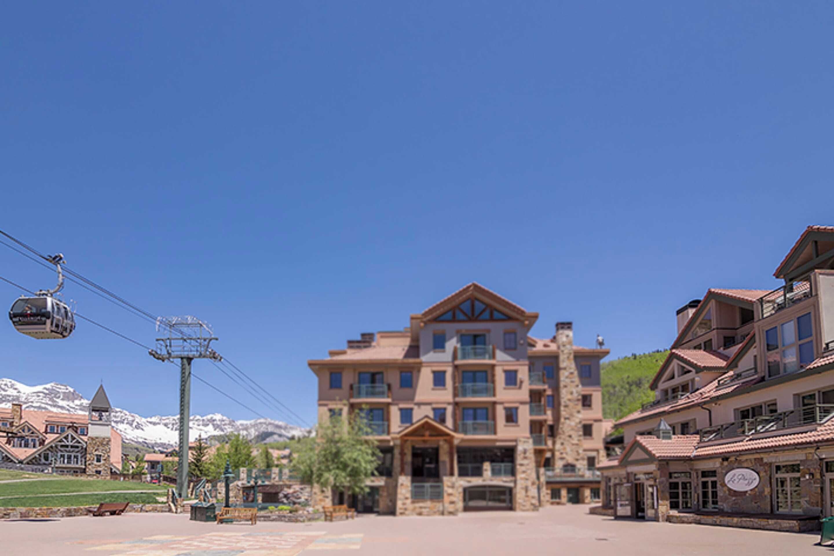 A multi-story hotel with a stone facade, a ski lift in the background, and a clear blue sky.