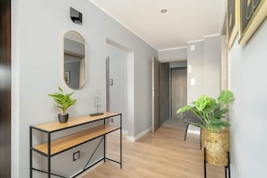 Un pasillo con piso de madera, un escritorio con planta en maceta, un espejo fijado a la pared y una puerta que da a otra habitación.