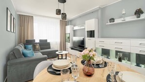 Un salon moderne avec un canapé gris, une table à manger dressée avec des assiettes et des verres, et un coin cuisine doté de meubles blancs et d’un téléviseur.