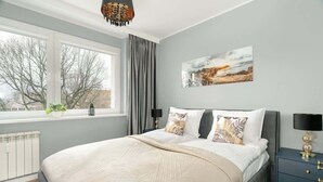 Une chambre avec un grand lit, une fenêtre donnant sur des arbres, un radiateur et une table de chevet avec une lampe.