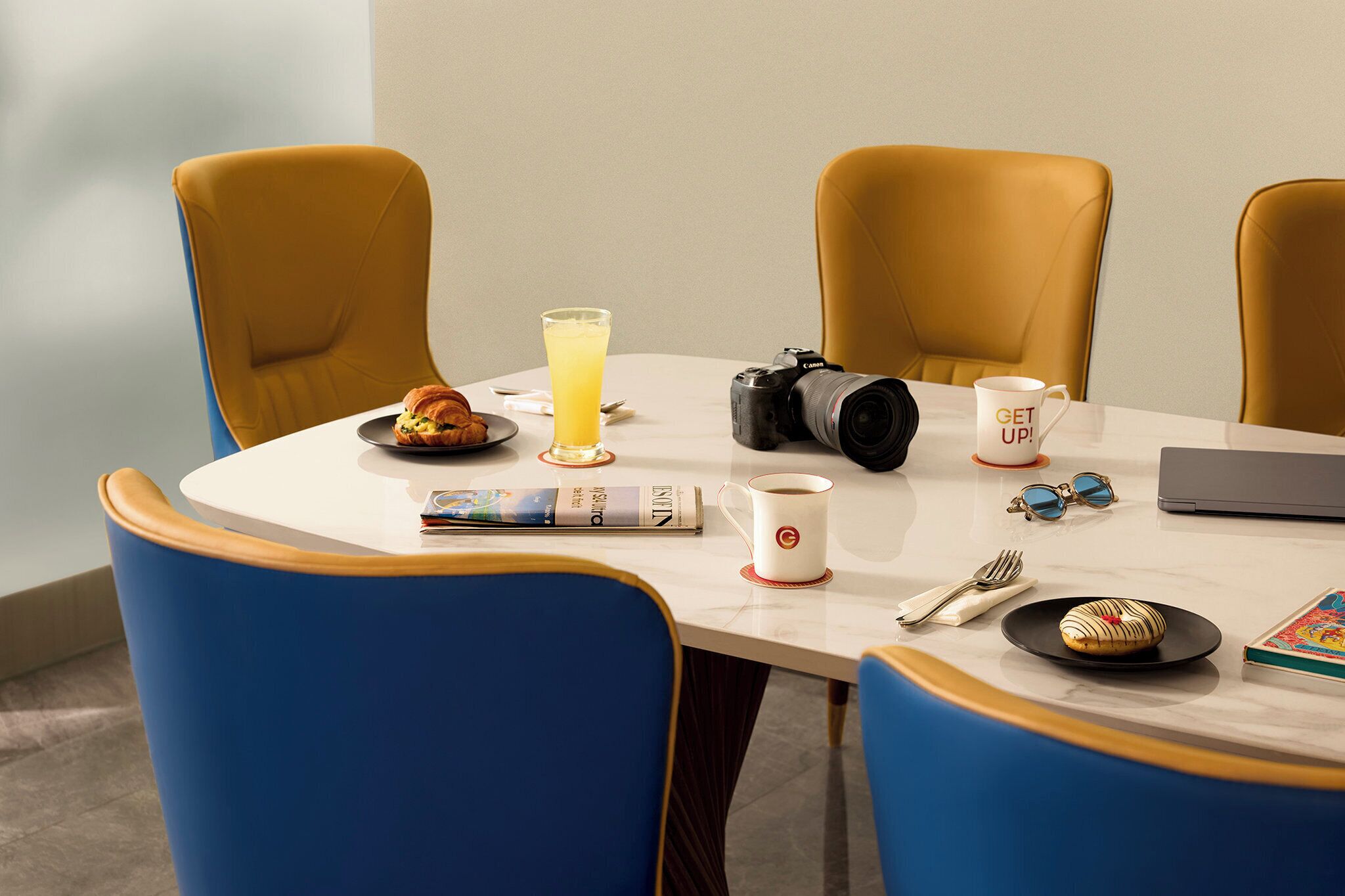 Una mesa de conferencias con una cámara, un vaso de jugo de naranja, un croissant, una taza, un par de gafas de sol, un libro y una computadora portátil.