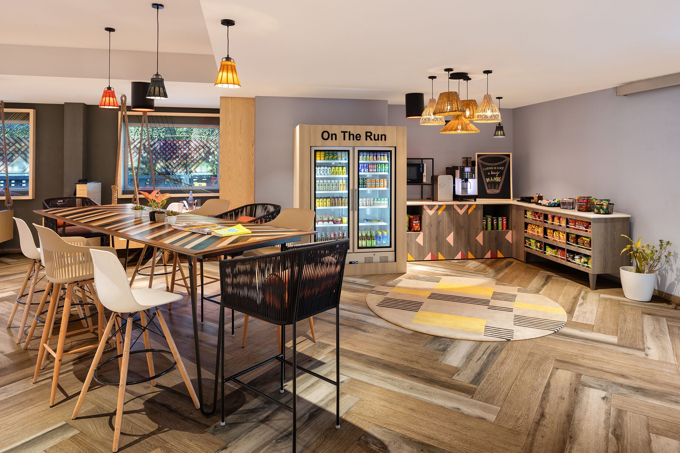Ein modernes Café mit Holztisch und -stühlen, einem Kühlschrank mit der Aufschrift 'On The Run' und einer Auswahl an Snacks und Getränken.
