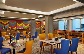 Un restaurant moderne aux murs ornés d’un motif à chevrons éclatant, avec des chaises jaunes et bleues, et une variété de plats présentés sur des étagères.