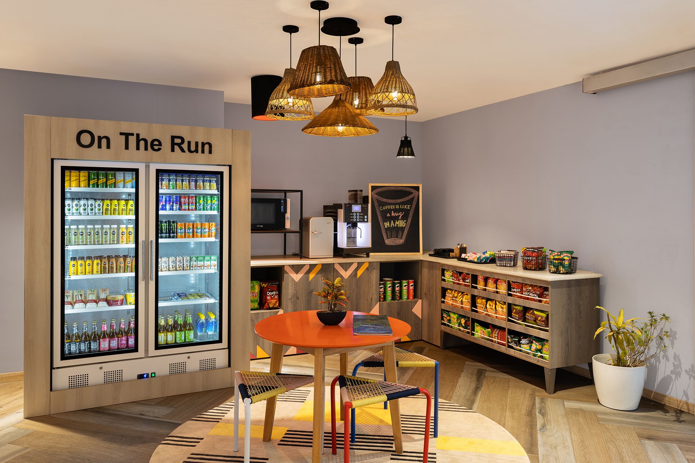 A modern room with a "On The Run" refrigerator stocked with beverages, a coffee machine, a dining table with chairs, and a bookshelf with snacks.
