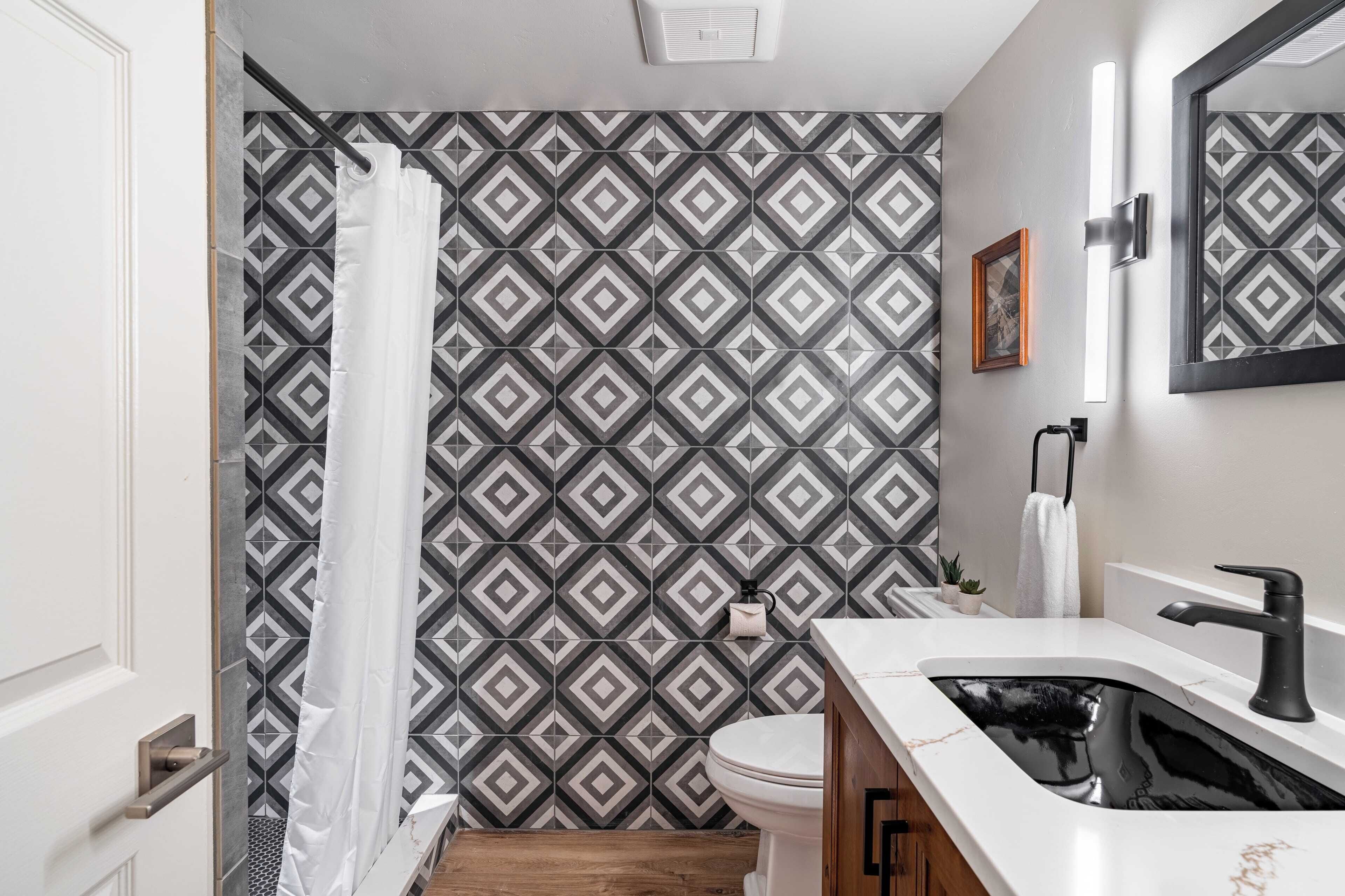 Une salle de bain avec un mur recouvert de carreaux décoratifs, des toilettes blanches, un lavabo noir avec un miroir et un rideau de douche.