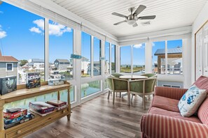 Un solarium con ventanas amplias, un ventilador de techo, una mesa de madera con juegos de mesa, un sillón verde y un sofá a rayas.
