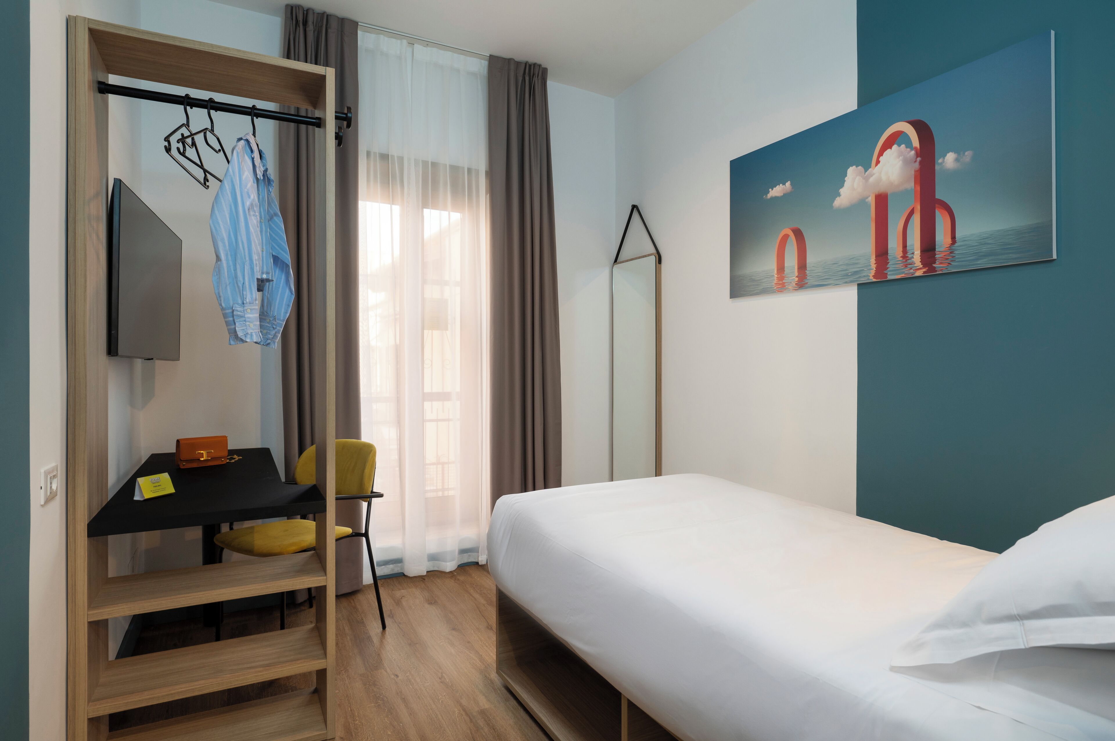 A hotel room with a bed, a desk with a chair, a TV, and a painting on the wall.