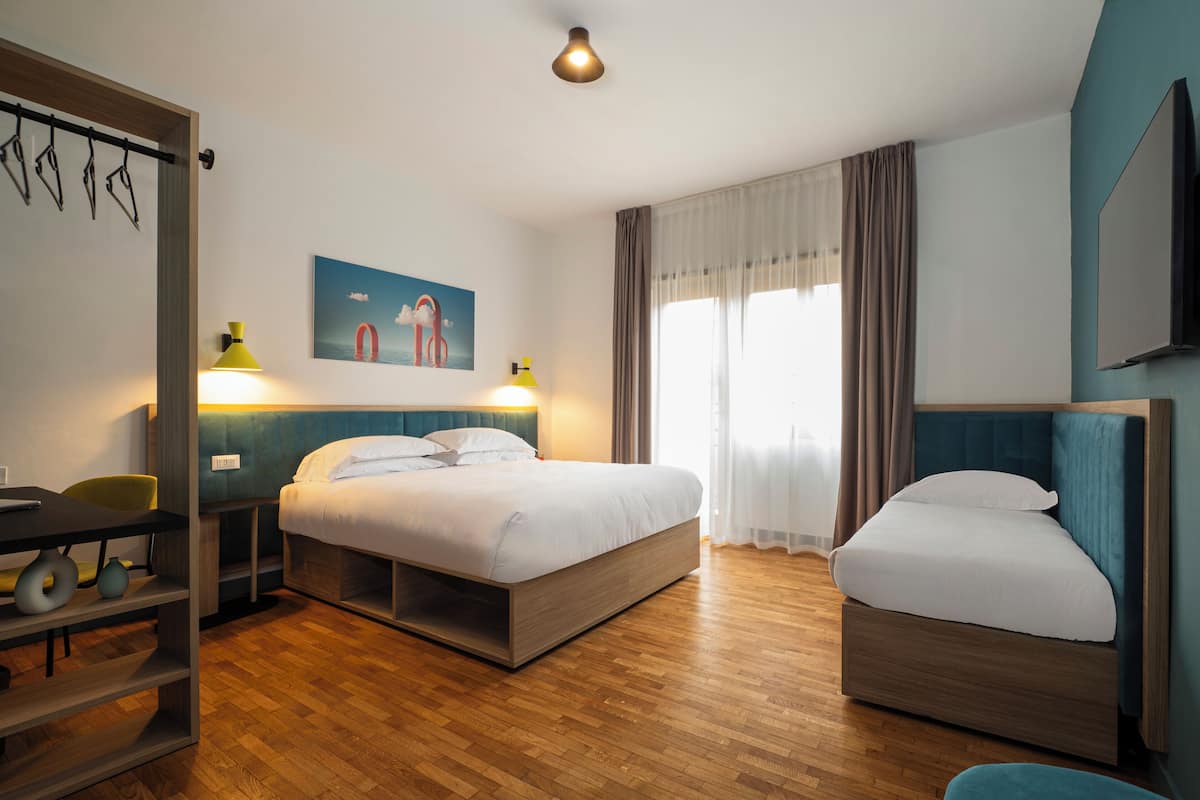 A modern hotel room with a wooden floor, a bed with a headboard, a desk with a chair, a TV, and a window with curtains.