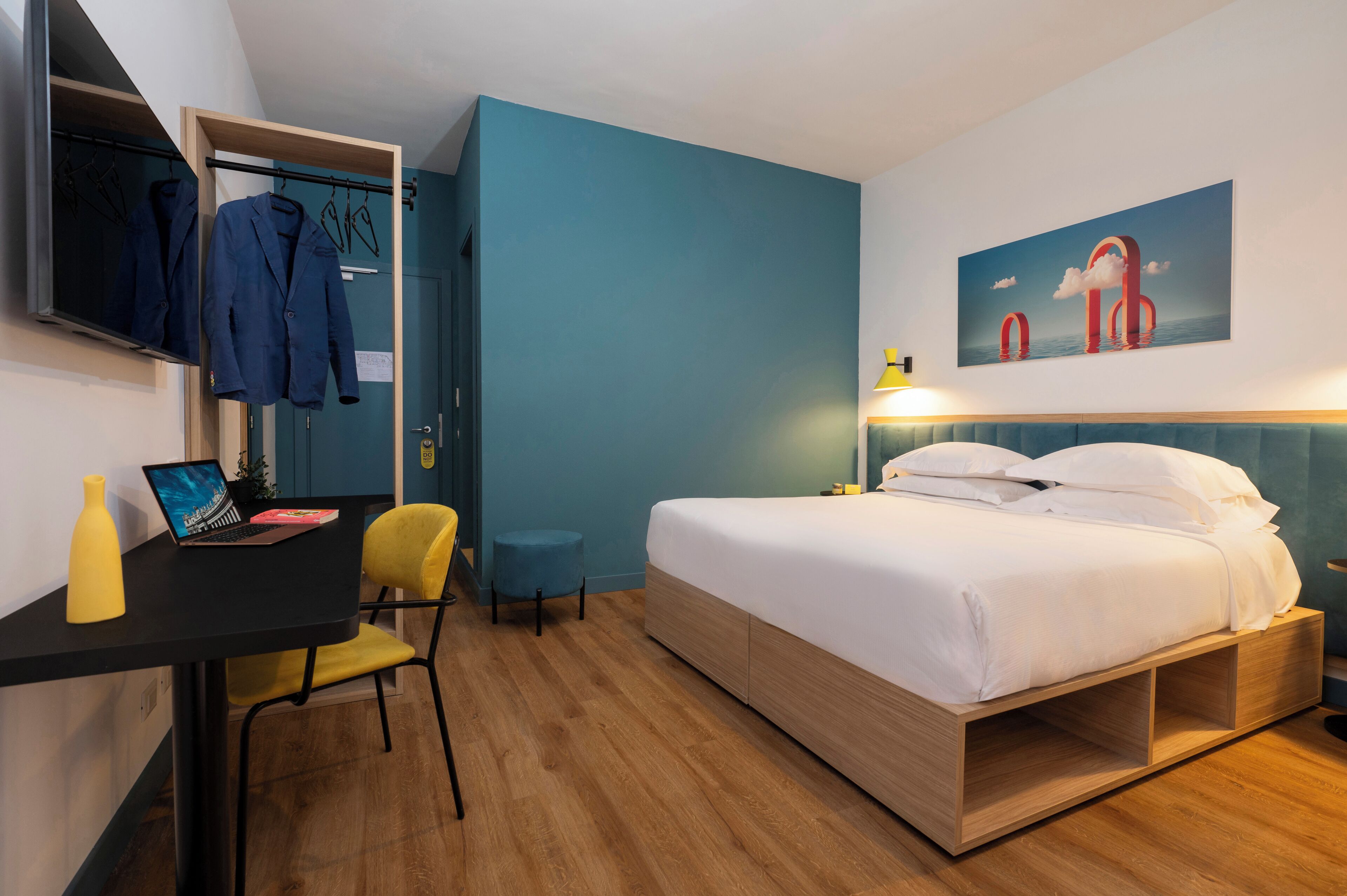 Ein modernes Hotelzimmer mit einem Bett, einem Schreibtisch mit einem Laptop, einem Sessel, einem Kleiderschrank und einem Gemälde an der Wand.