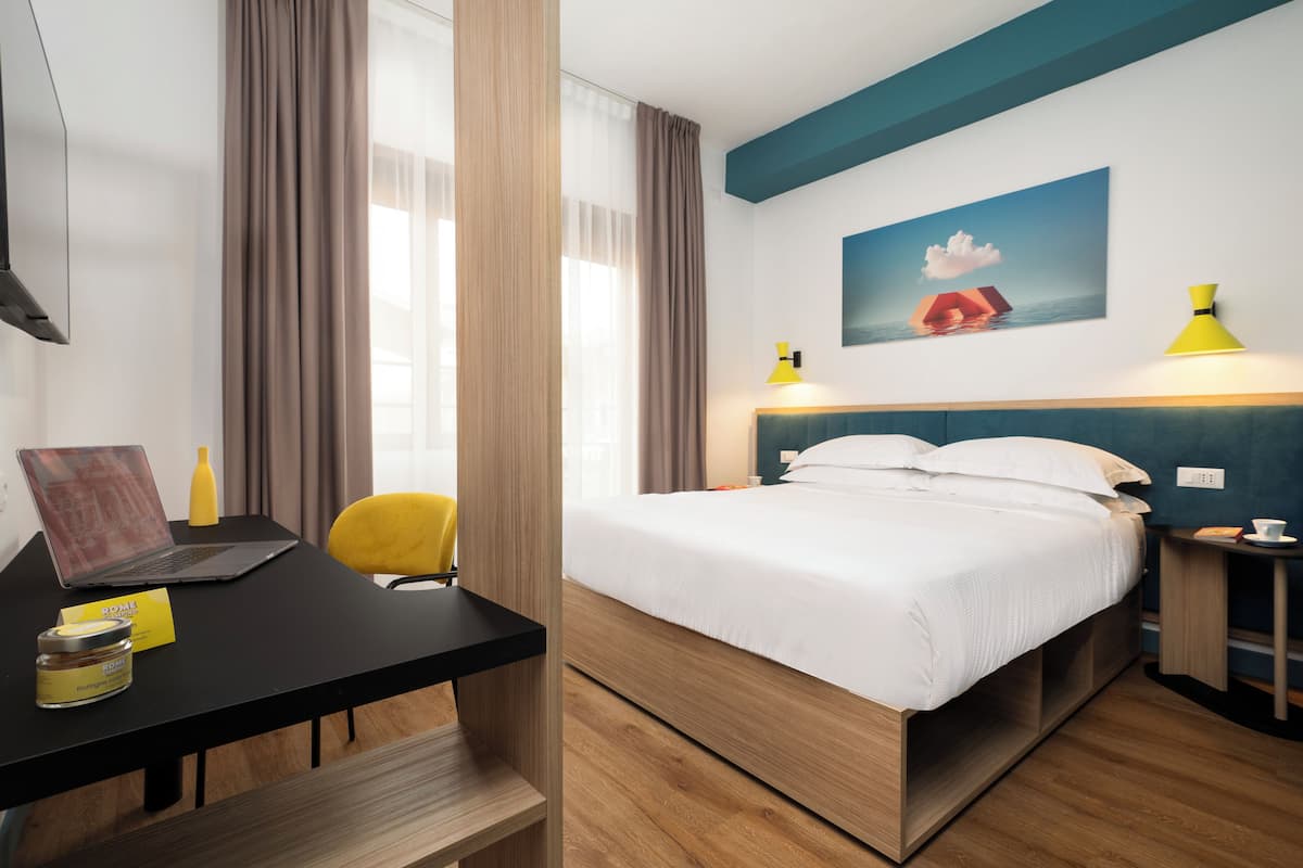 A hotel room with a bed, a desk with a laptop, a chair, a small table with a lamp, and a painting on the wall.