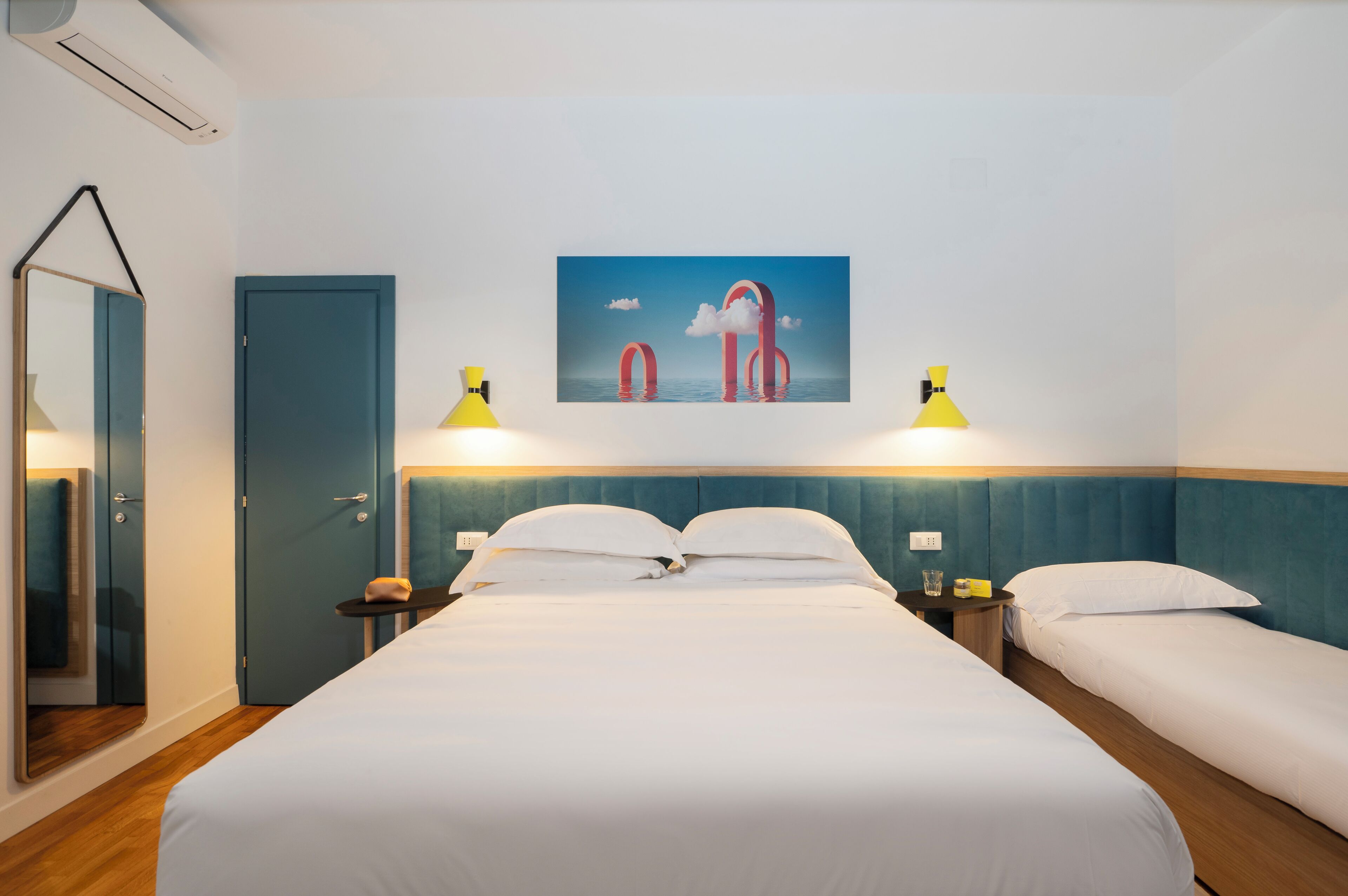 Habitación de hotel con una cama grande, dos mesitas de noche, un espejo, una puerta y un cuadro en la pared.