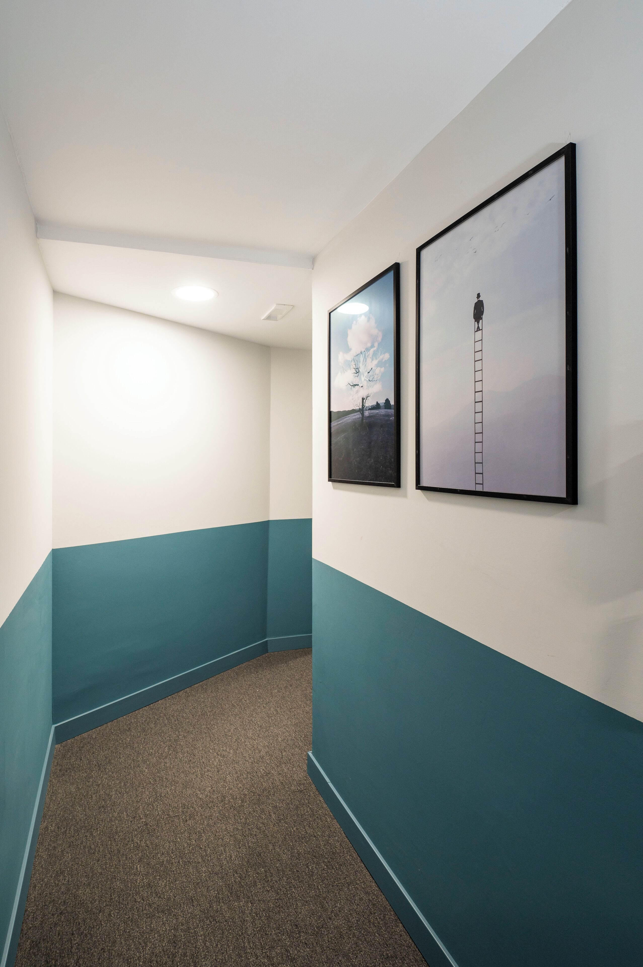 Un couloir aux murs de couleur turquoise et blanche, doté d’un sol recouvert de moquette et orné de deux tableaux encadrés.