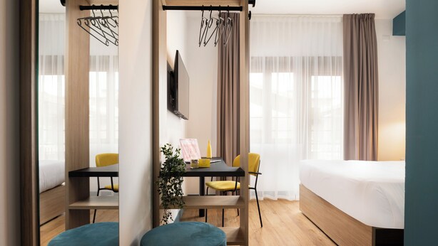 Une chambre d’hôtel moderne équipée d’un lit, d’un bureau avec des chaises, d’une télévision et d’une fenêtre avec des rideaux.