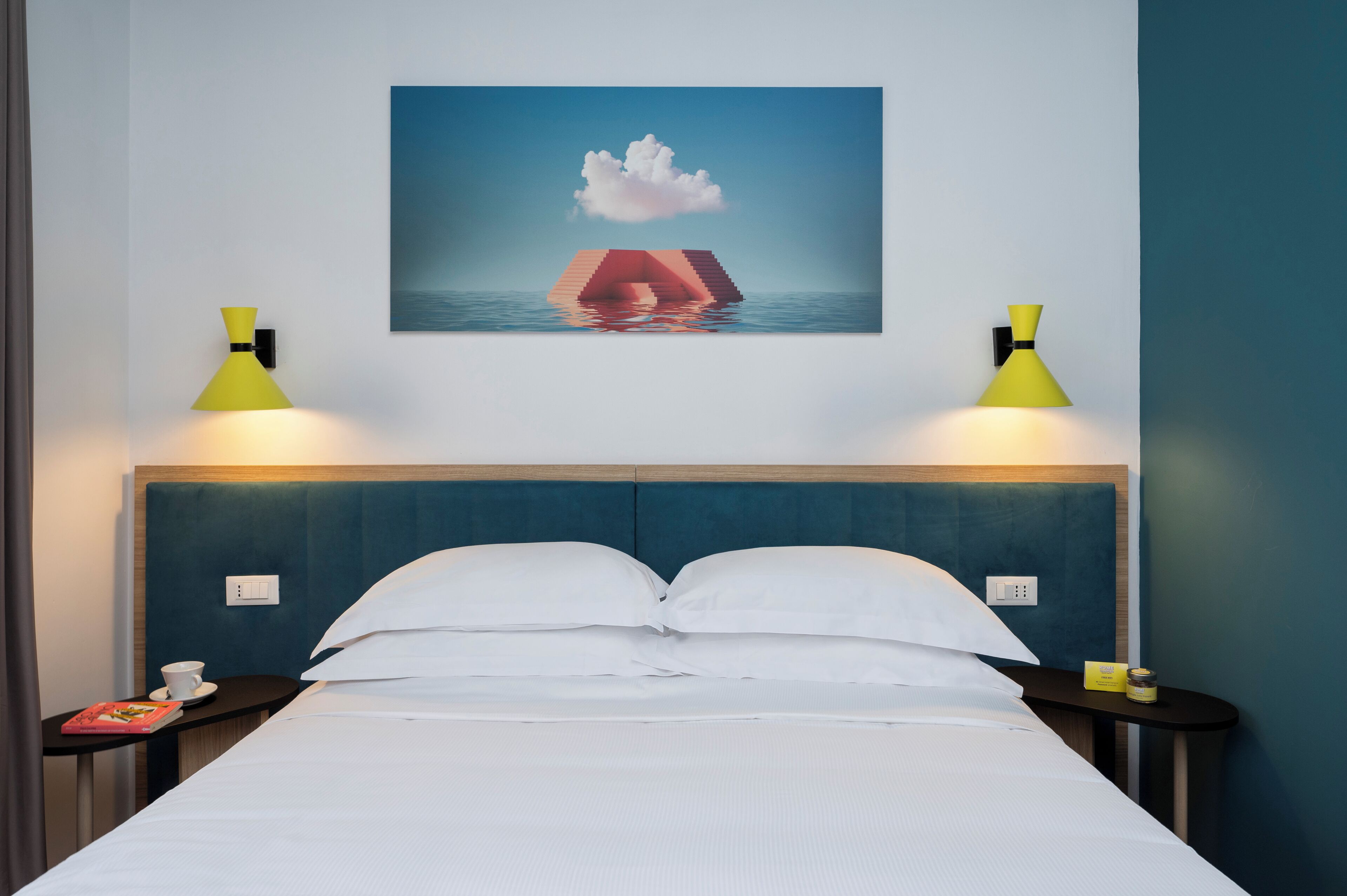 Une chambre d’hôtel avec un lit, deux tables de chevet, une lampe fixée au mur, un tableau représentant un nuage et une montagne, et une vue sur la mer.