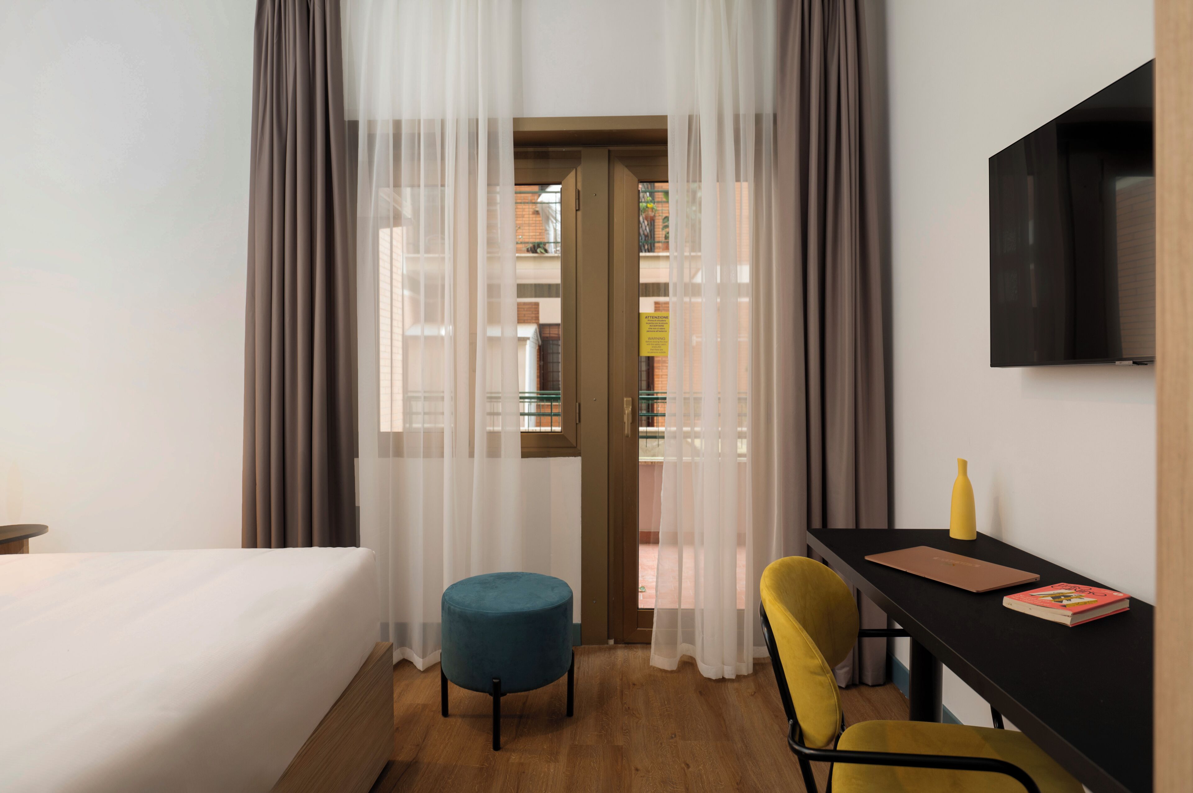 Habitación de hotel con cama, escritorio con silla, un pequeño taburete redondo, una silla amarilla, televisor montado en la pared y cortinas de gasa.
