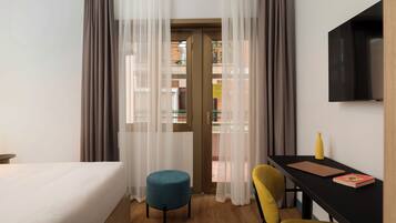 Habitación de hotel con cama, escritorio con silla, un pequeño taburete redondo, una silla amarilla, televisor montado en la pared y cortinas de gasa.