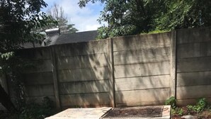 Une structure en béton surmontée d’une grille métallique, entourée d’une base en béton, située dans un jardin avec une clôture en bois et des arbres en arrière-plan.