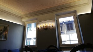 Un comedor con candelabro, dos ventanas con contraventanas y un techo con molduras decorativas.
