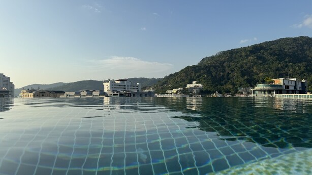 A waterfront hotel with a pool, buildings, and a forested hill in the background. - Zephyr Grand Hotel (Patong)
