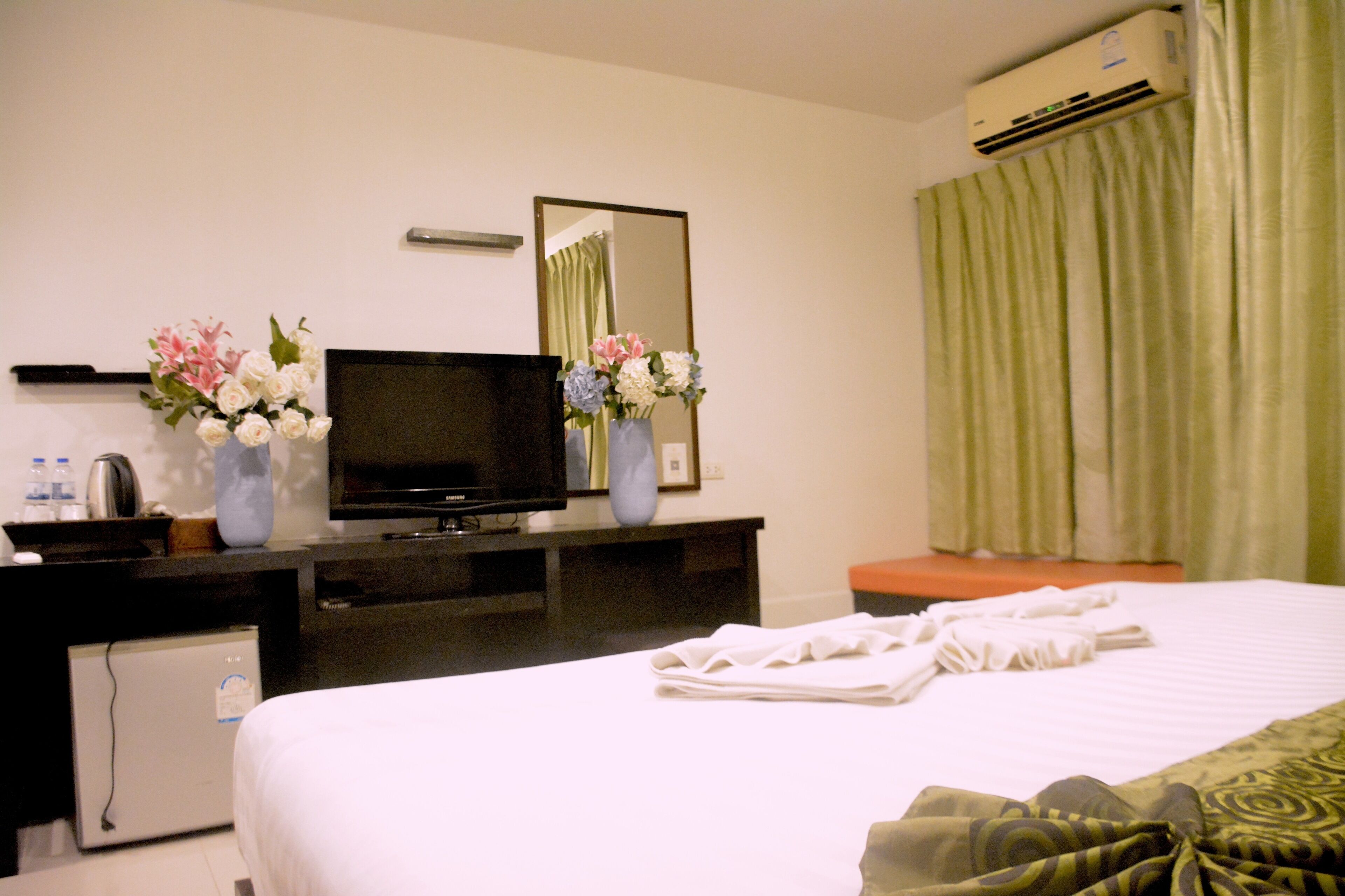 A hotel room with a bed, a television, a small refrigerator, a desk with a mirror, and two flower arrangements.