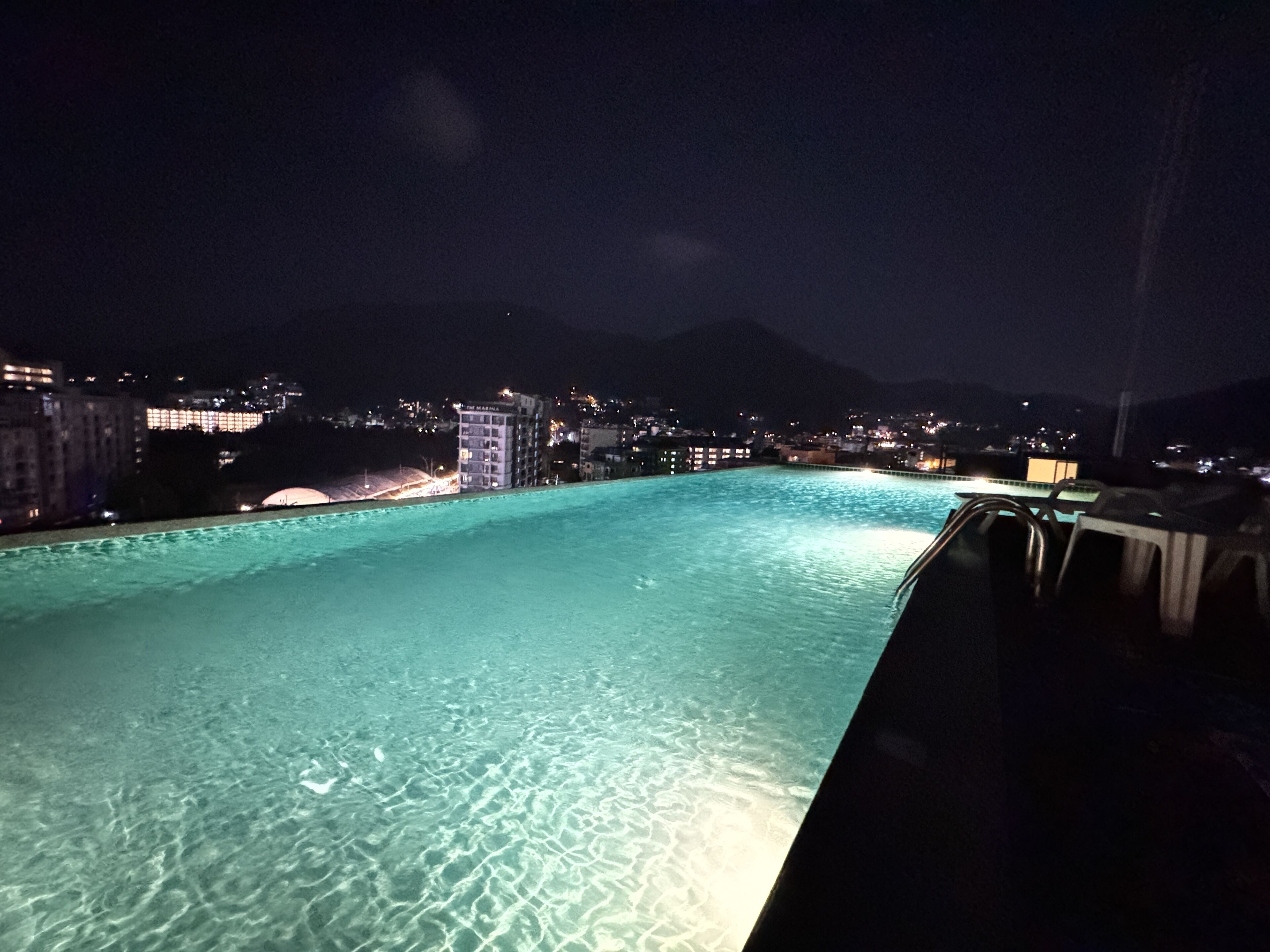 A nighttime view of a swimming pool with clear water, surrounded by buildings and a mountain in the background.