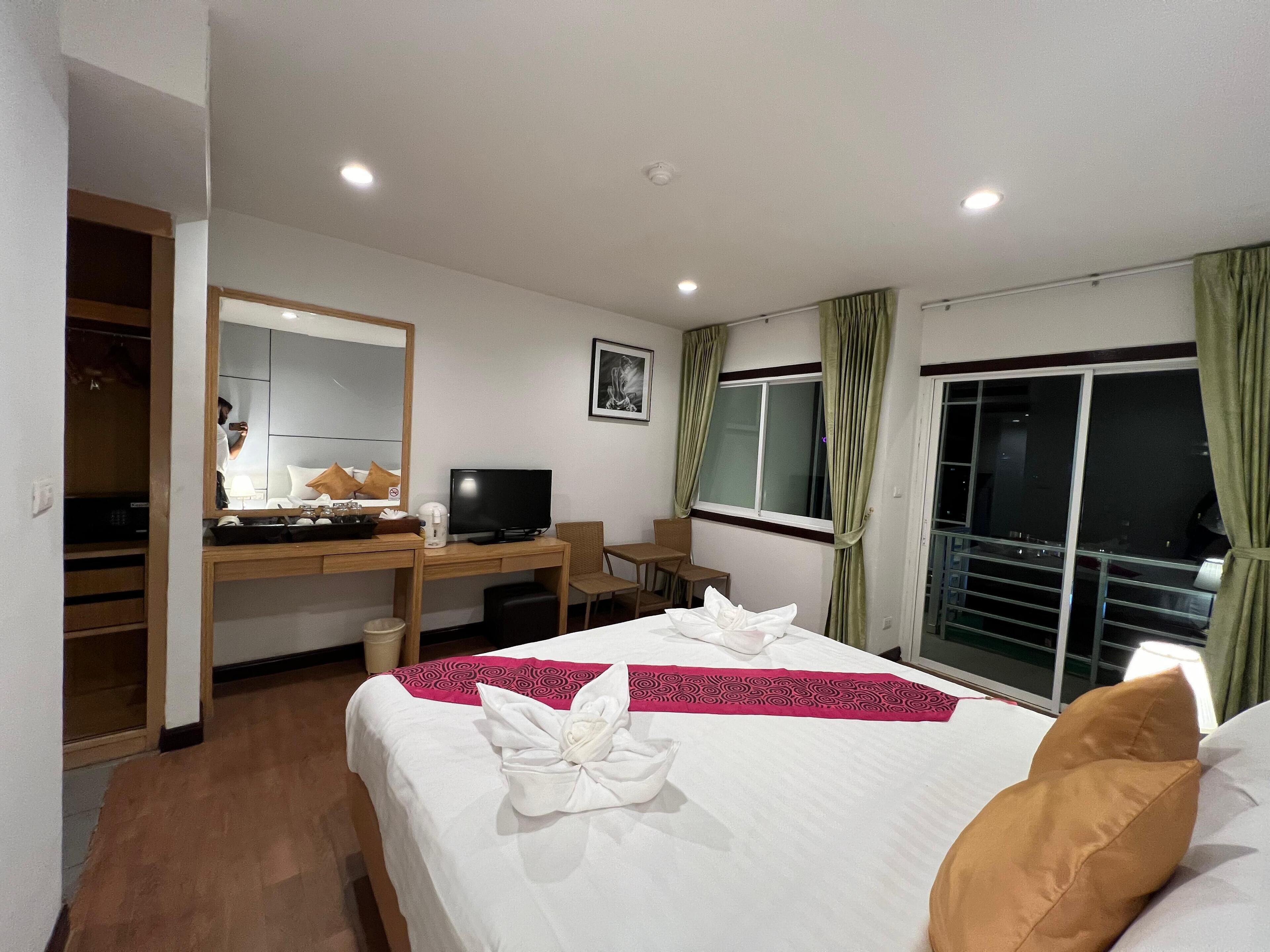 A hotel room with a bed, a desk with a computer, a chair, a window with curtains, and a balcony.